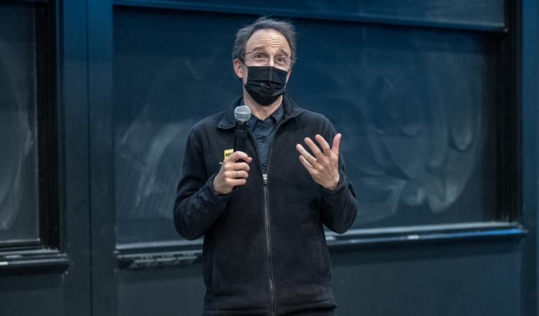 Masked professor stands in front of chalkboard holding microphone